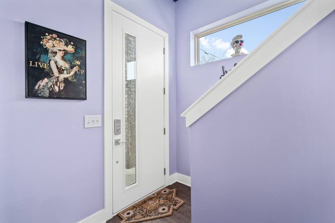 Bright lavender residential entryway with a modern white front door featuring a vertical glass insert, patterned doormat on dark hardwood floor, vintage floral portrait on the wall, and a playful decorative bust peeking through the stair window.