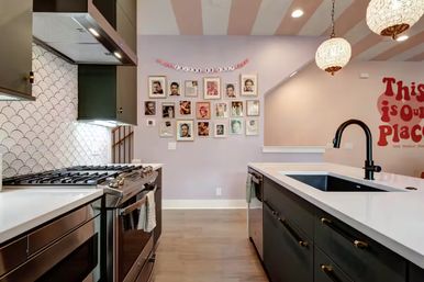 Modern pastel kitchen with white quartz island and black faucet, stainless steel gas range, scallop tile backsplash, framed gallery wall, pink-striped ceiling and pendant lights.