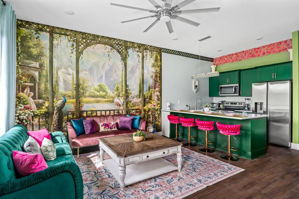 Vibrant open-concept living room and kitchen with jewel-toned sofas, scenic peacock and mountain wall mural, emerald green cabinets, hot-pink bar stools, rustic coffee table and hardwood floors
