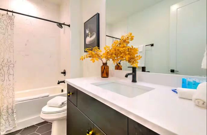 Modern bathroom interior with white quartz countertop and undermount sink, matte black faucet and vanity, bathtub with shower and patterned curtain, dark hexagon tile floor, large mirror and amber vase of yellow branches.