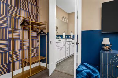 Chic navy-and-gold dressing area with geometric wallpaper, gold open clothing rack and quilted blue ottoman, opening through a white door to a modern bathroom with white double-sink vanity and large mirror.