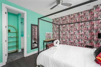 Bright guest bedroom with turquoise walls and pink cactus wallpaper, black four‑poster bed with white linens and towel swan, lighted vanity mirror, ceiling fan and open doorway to a bathroom.