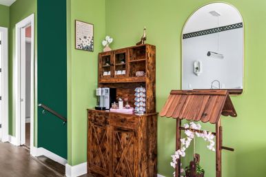 Bright green home hallway with rustic wooden hutch turned coffee station, K-cup carousel, arched wall mirror, and decorative wooden wishing-well planter with pink blossoms