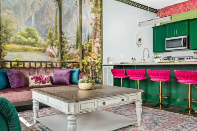 Bright, eclectic open-plan living room and kitchen with emerald green cabinets, four hot-pink velvet bar stools, mauve sofa with jewel-tone pillows, distressed white coffee table, floral rug, and a scenic garden mural with a swan and lake.