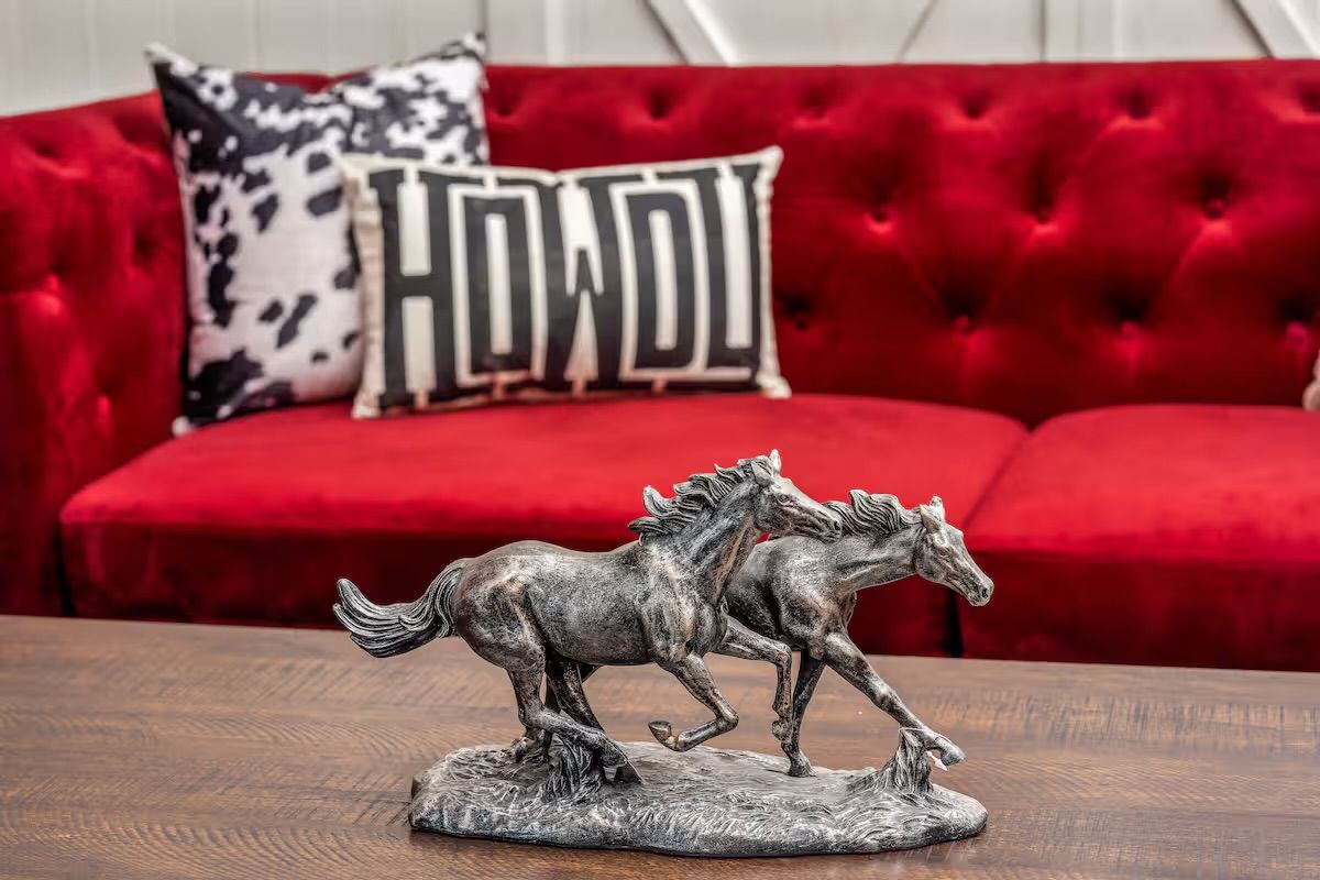 Two galloping bronze horse sculptures on a wooden coffee table in front of a tufted red velvet sofa with a black-and-white "HOWDY" pillow and cowhide accent pillow — rustic southwestern living room decor.