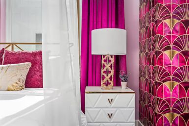 Luxury bedroom nook with fuchsia velvet pillows, magenta curtains, gold art-deco wallpaper, white bedside table, ornate gold lamp and small potted plant