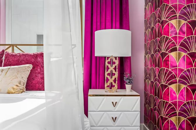 Luxury bedroom nook with fuchsia velvet pillows, magenta curtains, gold art-deco wallpaper, white bedside table, ornate gold lamp and small potted plant