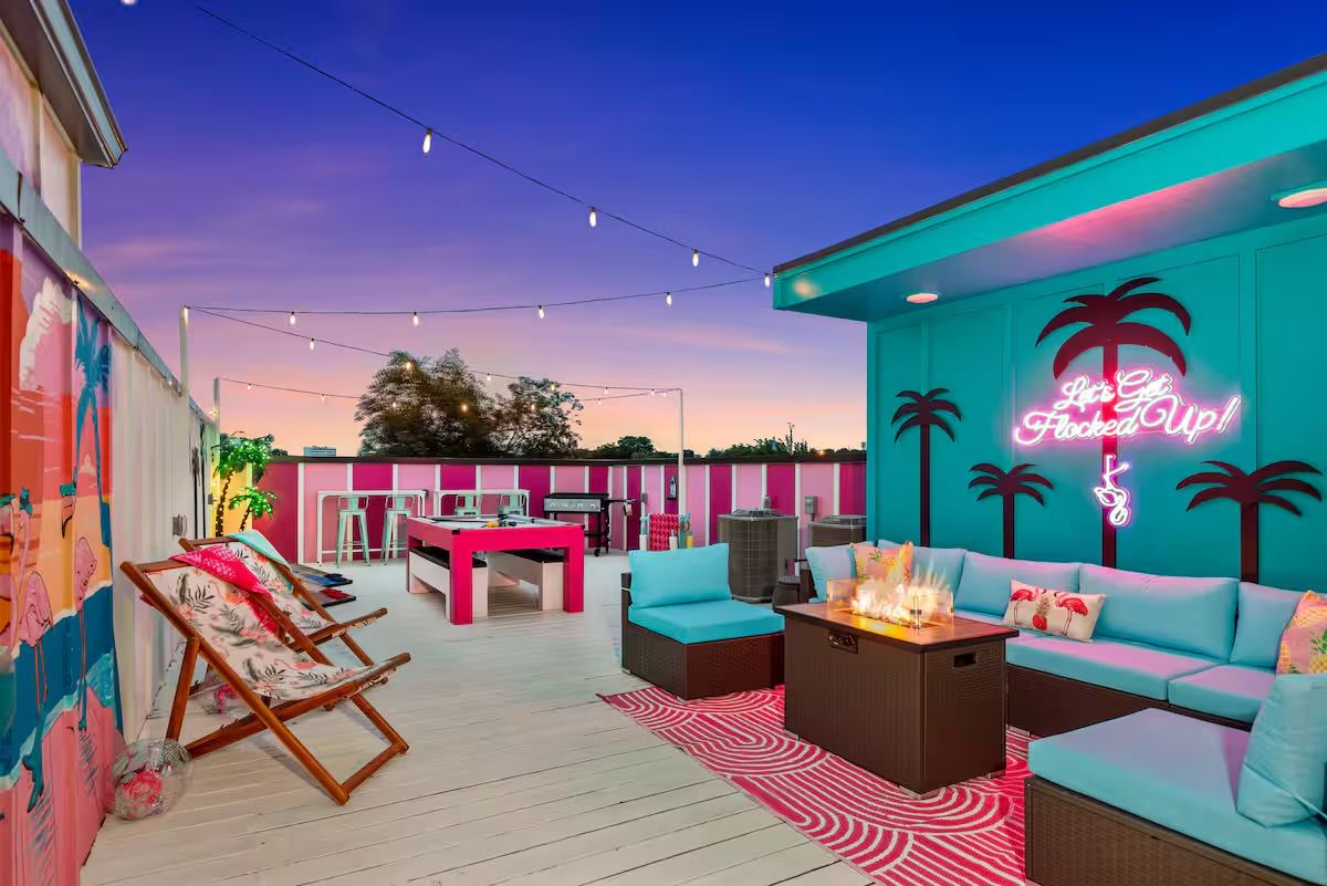 Vibrant rooftop lounge at sunset with teal and pink tropical décor, string lights and neon palm sign, turquoise sectional seating around a fire table, lounge chairs and high-top tables.