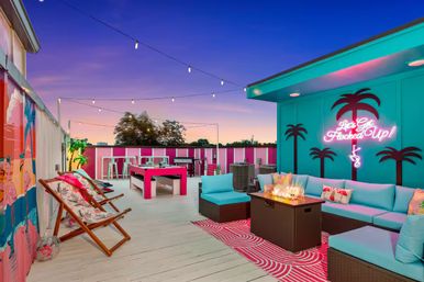 Vibrant rooftop lounge at sunset with teal and pink tropical décor, string lights and neon palm sign, turquoise sectional seating around a fire table, lounge chairs and high-top tables.