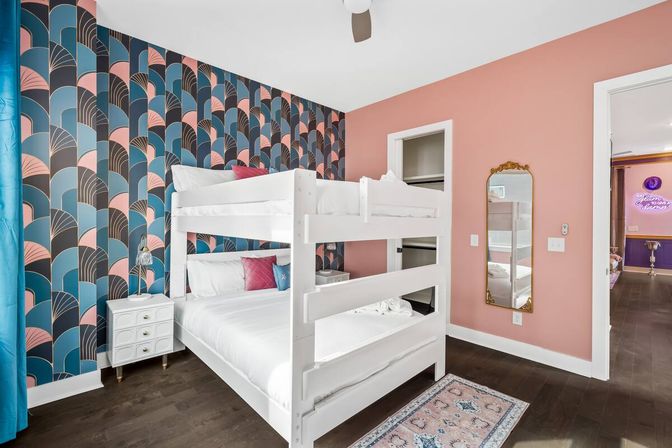 Playful modern bedroom with a white double bunk bed, teal-and-pink art-deco accent wallpaper, coral painted wall, dark hardwood floors, white nightstand, gold-framed mirror and patterned rug.