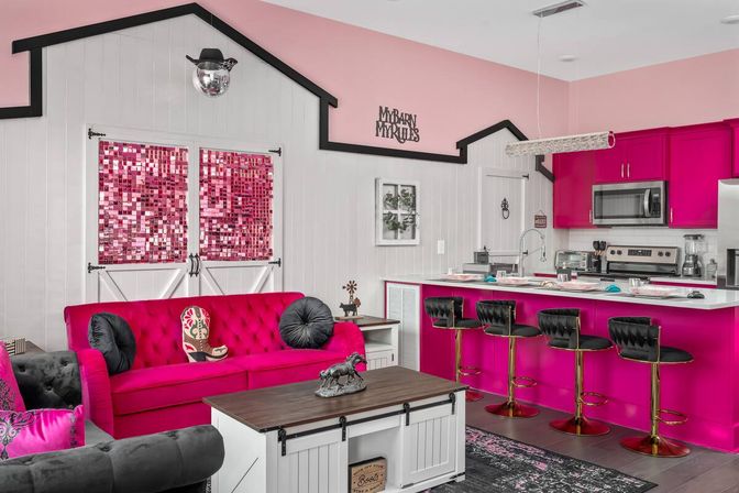 Open-plan farmhouse-style living room and kitchen with hot-pink tufted sofa, magenta cabinets, white shiplap walls, barn doors with pink sequin panels, four black bar stools, horse figurine and disco-ball with cowboy hat.
