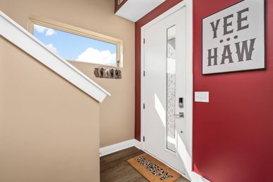 Bright western-themed entryway with a white front door and vertical glass panel, red accent wall with 'YEE HAW' sign, 'Howdy' doormat on hardwood floor, tan stair wall and narrow sky window.