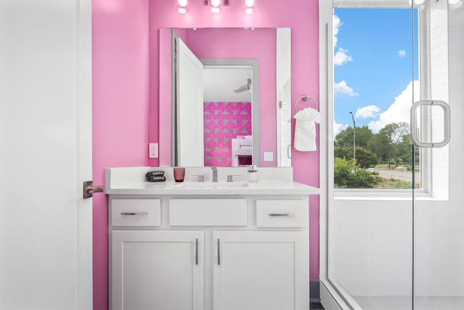 Playful bright-pink bathroom with a white vanity and marble countertop, illuminated mirror and bulbs, towel ring, glass shower door and a large window showing a tree-lined street view.
