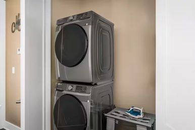 Stacked front‑load washer and dryer in a compact beige home laundry closet, with an iron resting on a nearby folding shelf.