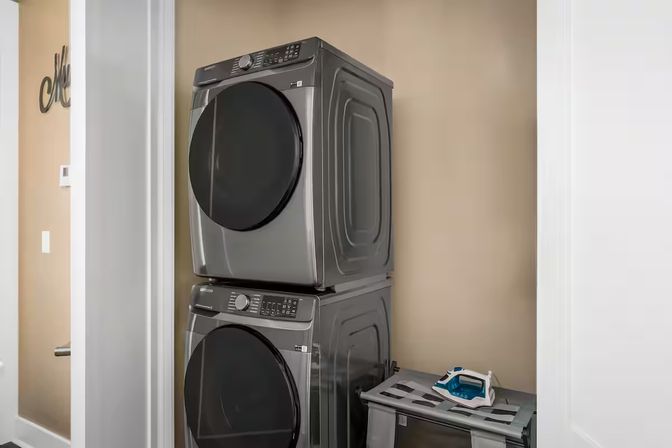 Stacked front‑load washer and dryer in a compact beige home laundry closet, with an iron resting on a nearby folding shelf.