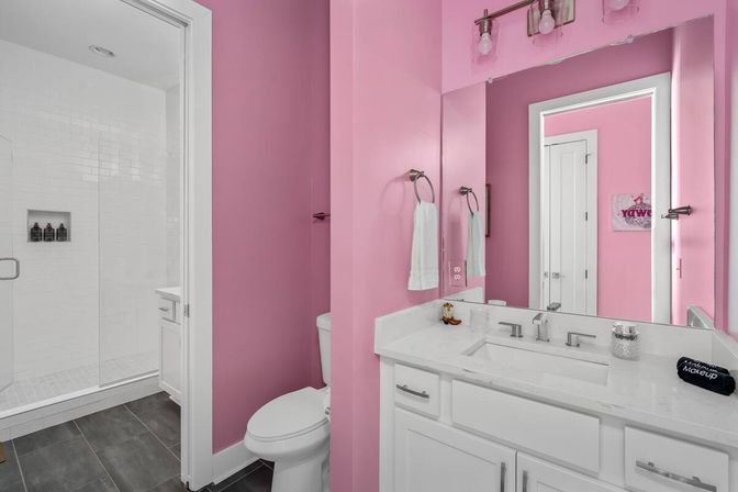 Playful pink bathroom with white vanity and quartz countertop, undermount sink and large mirror, glass-enclosed white subway-tile shower, white toilet, towel ring, and dark gray floor tiles.