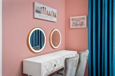 Stylish makeup vanity nook with pink walls, two round LED-lit mirrors above a white quilted desk, two gray velvet chairs with gold ring pulls and teal curtains
