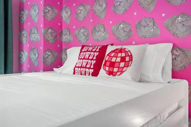 Playful bedroom interior with white platform bed, decorative red "HOWDY" pillow and red disco-ball pillow, set against bold pink wallpaper patterned with sketched cowboy hats and stars.