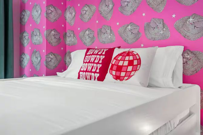 Playful bedroom interior with white platform bed, decorative red "HOWDY" pillow and red disco-ball pillow, set against bold pink wallpaper patterned with sketched cowboy hats and stars.