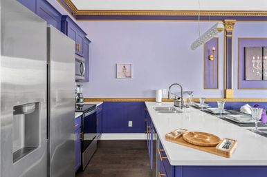 Luxury purple kitchen with lavender walls and gold moldings, white quartz island with sink and wooden serving tray, stainless steel refrigerator and oven, crystal glassware and a linear pendant light