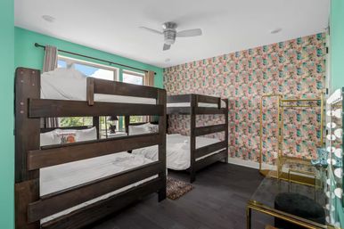 Coastal-themed bedroom with two dark wooden bunk beds (four beds), mint-turquoise walls, colorful tropical wallpaper, large windows, ceiling fan, gold clothing rack and lighted vanity on dark hardwood floors.
