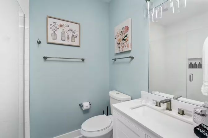Bright, airy small modern bathroom with light-blue walls, white vanity and sink, toilet, chrome fixtures, floral wall art, and glass shower door