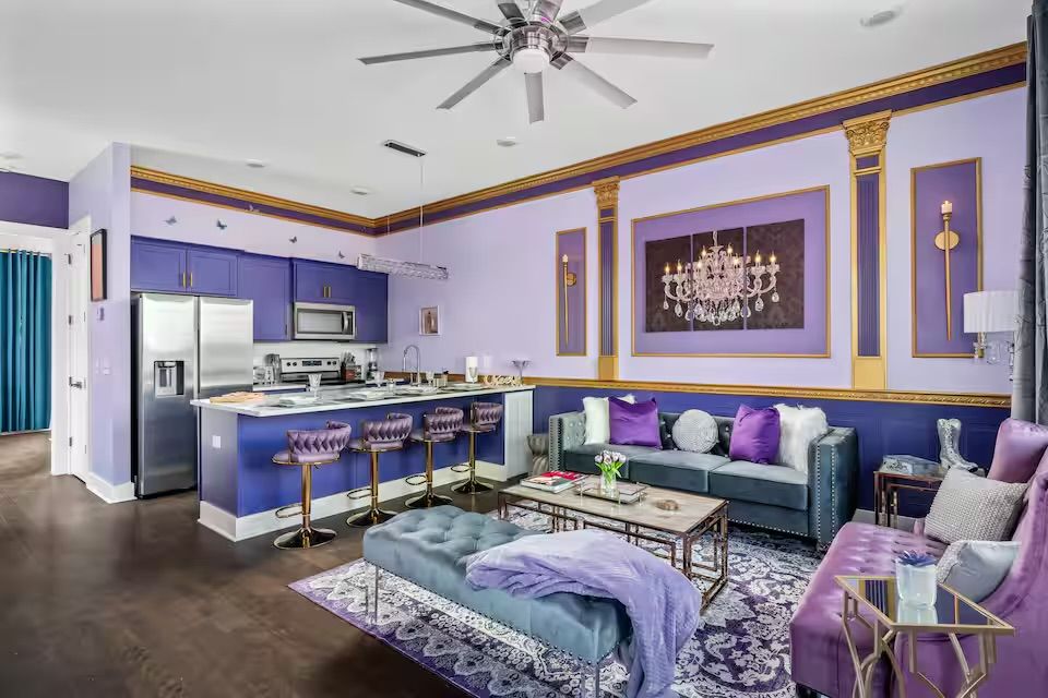 Regal purple-and-gold open-concept living room and kitchen with velvet seating, tufted bar stools, stainless appliances, marble coffee table and chandelier artwork