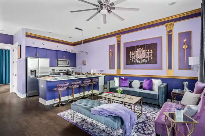Regal purple-and-gold open-concept living room and kitchen with velvet seating, tufted bar stools, stainless appliances, marble coffee table and chandelier artwork