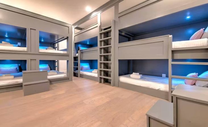 Modern sleep room with built-in gray bunk beds, white linens, blue LED accent lighting and light wood floors — cozy dorm-style bunk room for groups.