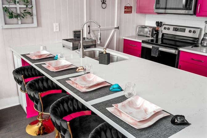 Bright modern kitchen island with white quartz countertop and stainless gooseneck faucet, hot-pink cabinets, four black velvet swivel bar stools with gold bases, and four place settings with pink-marble plates on gray placemats.