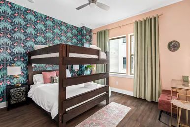 Modern urban bedroom with dark wood bunk bed, tropical teal flamingo-and-palm wallpaper, peach-painted walls, green curtains, ceiling fan and decorative nightstand
