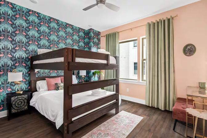Modern urban bedroom with dark wood bunk bed, tropical teal flamingo-and-palm wallpaper, peach-painted walls, green curtains, ceiling fan and decorative nightstand