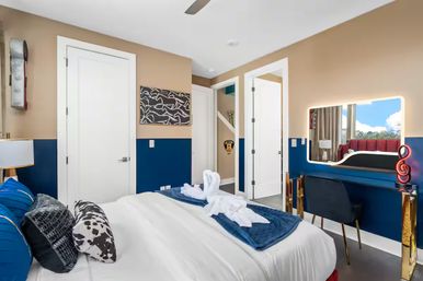 Bright modern bedroom with navy-and-beige half walls, white bed topped with towel swans and blue runner, illuminated vanity mirror with black chair and treble-clef accent, and a window view of blue sky and trees.
