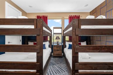 Bright family bunk room in a vacation rental with two dark-wood bunk beds facing each other, central nightstand and lamp under a window with red curtains, patterned accent wall, white linens and rolled towels — perfect for kids or groups.