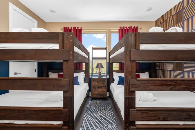 Bright family bunk room in a vacation rental with two dark-wood bunk beds facing each other, central nightstand and lamp under a window with red curtains, patterned accent wall, white linens and rolled towels — perfect for kids or groups.