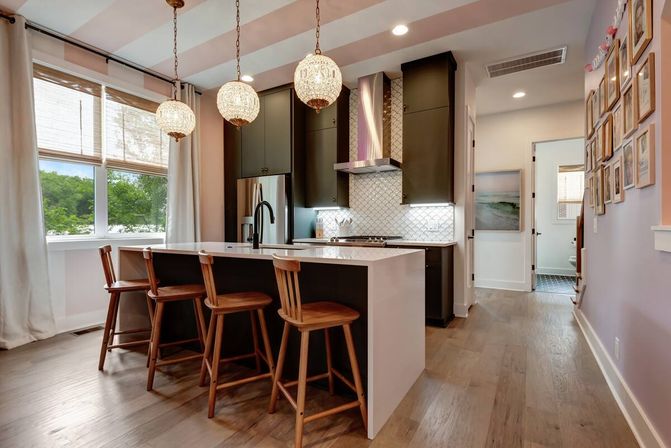 Bright modern kitchen with white waterfall island and three wooden bar stools, dark cabinets, stainless appliances, patterned tile backsplash, globe pendant lights, hardwood floors, and a large window with curtains.