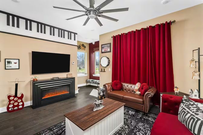 Bright modern living room with bold red curtains, tufted brown leather sofa, wall-mounted flat-screen TV above an electric fireplace, wooden coffee table, piano-key wall decal, decorative guitar and oversized ceiling fan.