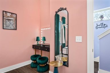 Pink dressing nook with black makeup vanity and lighted mirror, emerald green velvet poufs, tall ornate arched mirror reflecting window with green curtains, gold side table and framed wall art