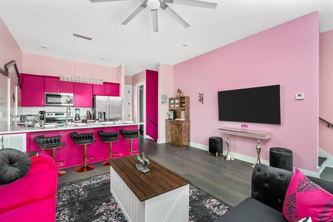 Bold pink open-concept living room and kitchen with hot-pink cabinets, white island with black bar stools, wall-mounted TV, gray hardwood floors, plush velvet seating and stainless steel appliances