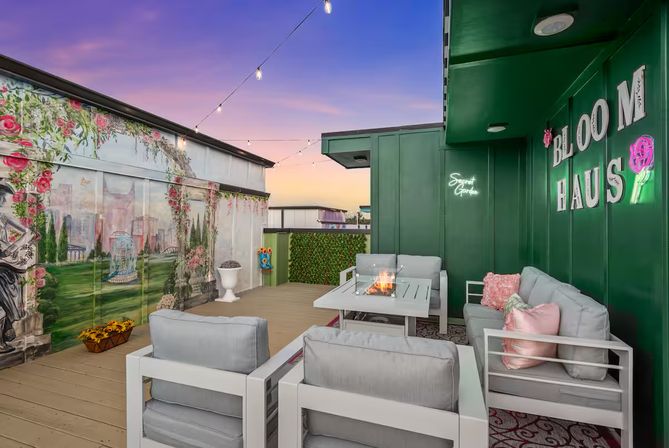 Urban rooftop terrace at sunset with string lights, colorful floral mural, green accent wall with neon garden sign, gray cushioned seating clustered around a fire pit table, pink accent pillows and potted decor.