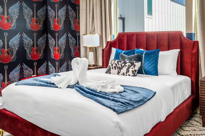 Rock-themed boutique hotel room with a red velvet tufted headboard, crisp white linens, blue throw and towel swans on the bed, blue and patterned accent pillows, and bold black wallpaper featuring red guitars with wings.