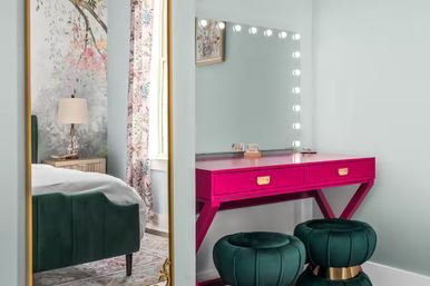 Hot-pink vanity table with lighted mirror, two emerald velvet poufs with gold bases, floral curtains and a reflected green upholstered bed in a chic bedroom corner.