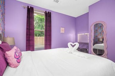 Playful purple bedroom with white bed topped by towel swans forming a heart, plum velvet curtains over a backyard window, illuminated vanity mirror and wavy full-length mirror