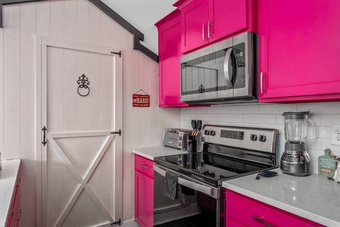 Bright modern kitchen with hot-pink cabinets, stainless-steel microwave and range, white shiplap barn-style door, toaster and blender on white countertops.