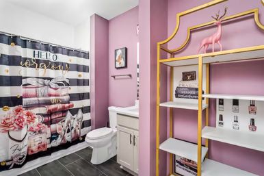 Pink home bathroom with ‘Hello Gorgeous’ striped shower curtain, white vanity and toilet, gold-trim open shelving displaying decorative books, nail polish bottles and a pink deer figurine, and dark tile floor.