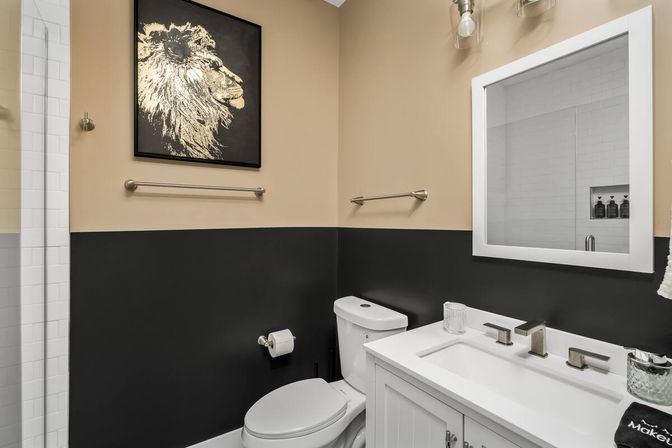 Modern small bathroom with white vanity and sink, white toilet, black lower half-wall and beige upper wall, framed gold lion artwork, mirrored medicine cabinet, and glass subway-tile shower visible.