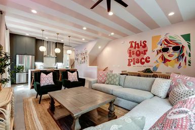 Open-plan living room and kitchen with pink-striped ceiling, colorful 'This is our place' mural, large light-gray sectional, green accent chairs, wooden coffee table, and pendant-lit kitchen island.