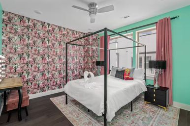Colorful modern bedroom interior with a black four-poster bed dressed in white linens and towel swans, pink cactus wallpaper, mint-green accent wall, pink curtains, vanity mirror lights and dark hardwood floor.