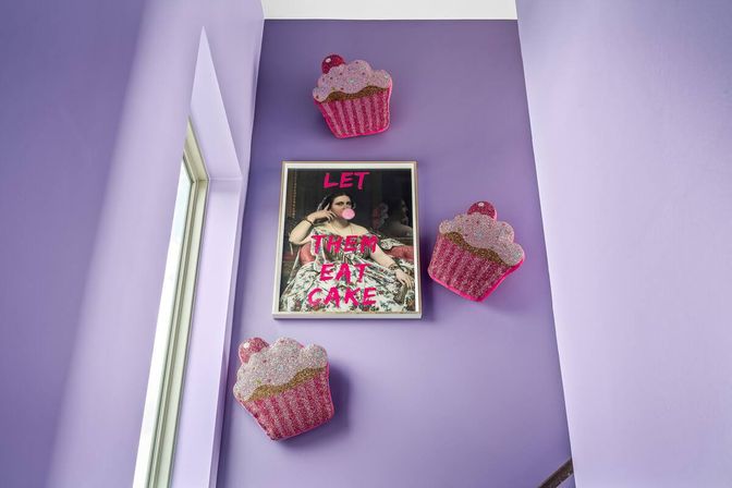Lavender stairwell with framed 'LET THEM EAT CAKE' print showing a woman blowing a pink bubble, flanked by three glittery pink cupcake wall sculptures.