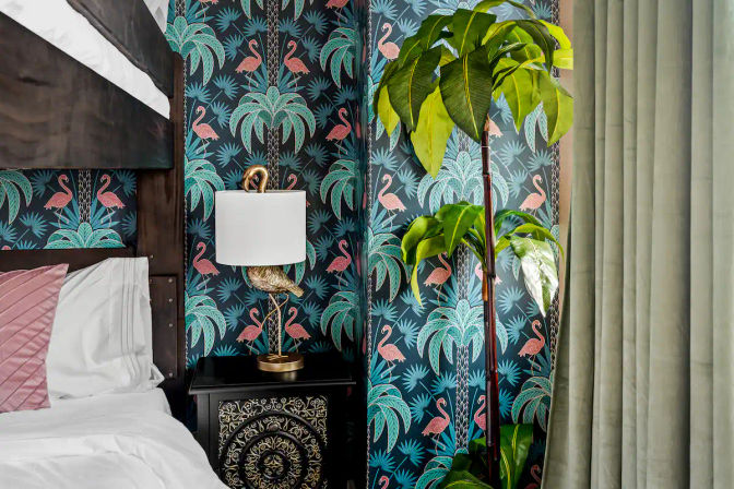 Playful tropical bedroom corner with teal flamingo-and-palm wallpaper, dark wood canopy bed, white bedding with a pink pillow, ornate black bedside table topped by a gold flamingo lamp, tall potted plant and green curtains.